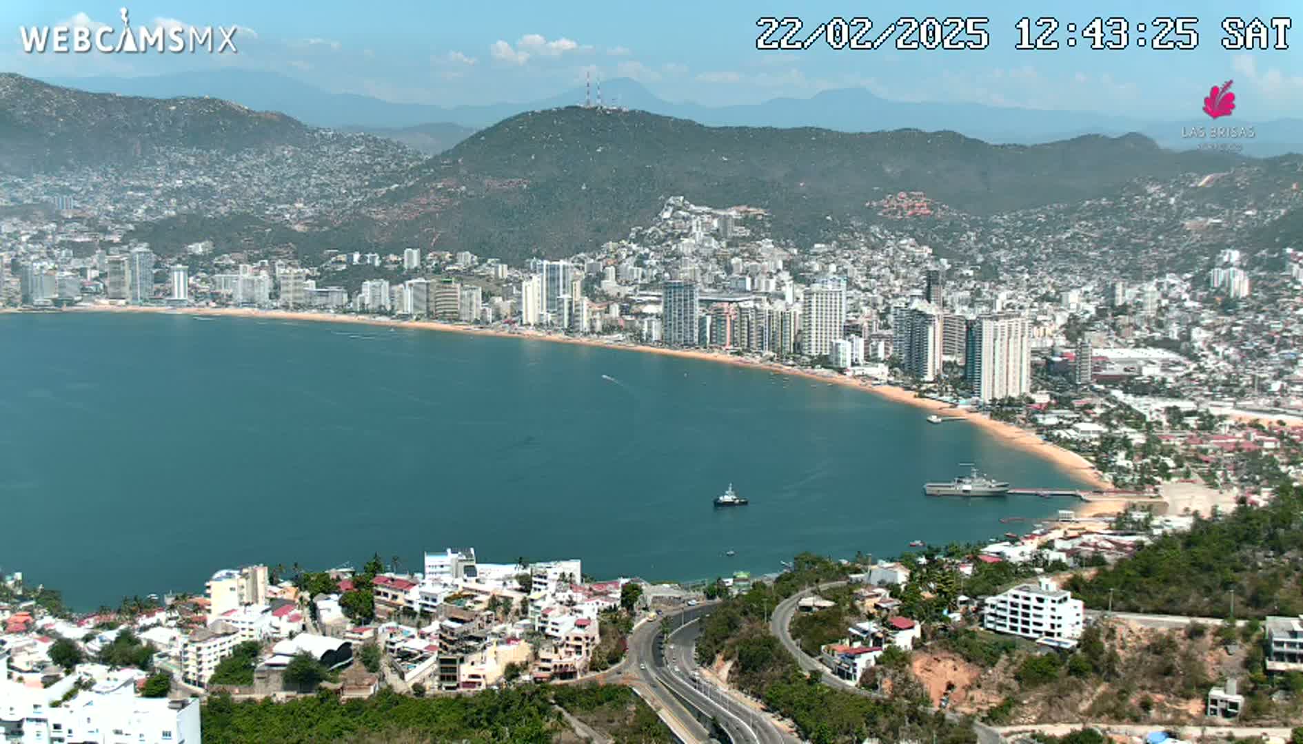 A coastal city with high-rise buildings and a sandy beach curves around a calm, teal bay under a mostly sunny sky.