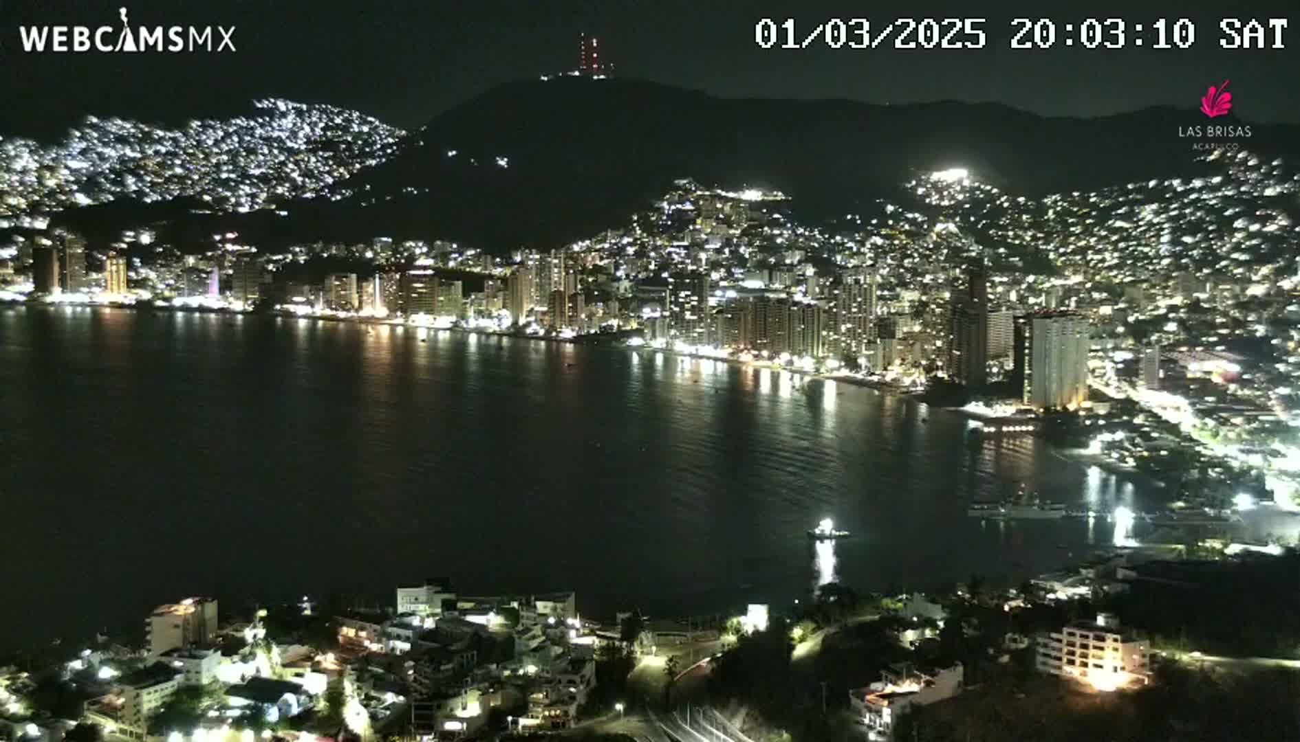 A nighttime view of a city and bay, illuminated by lights, with calm water reflecting the city lights.