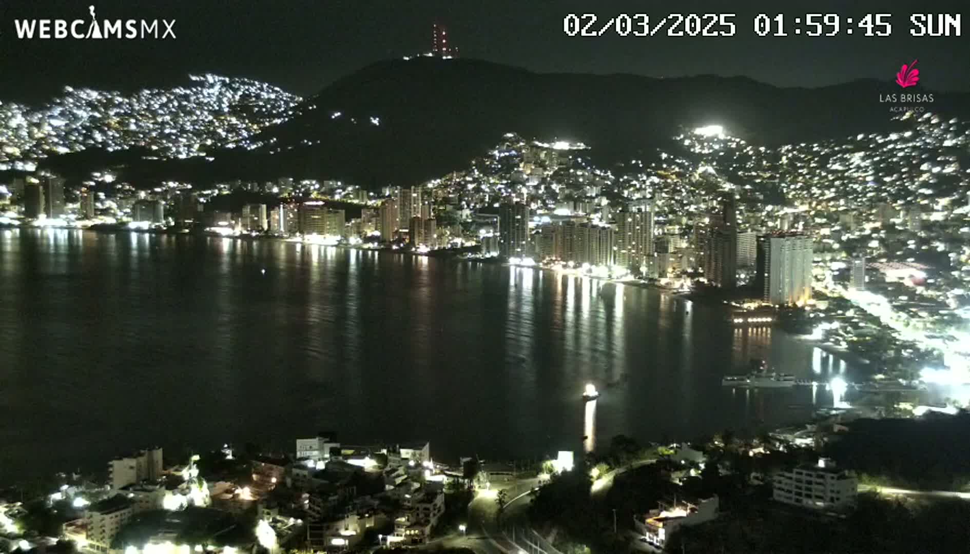 A nighttime view of a city illuminated along a dark bay, with hills rising behind it under a clear sky.