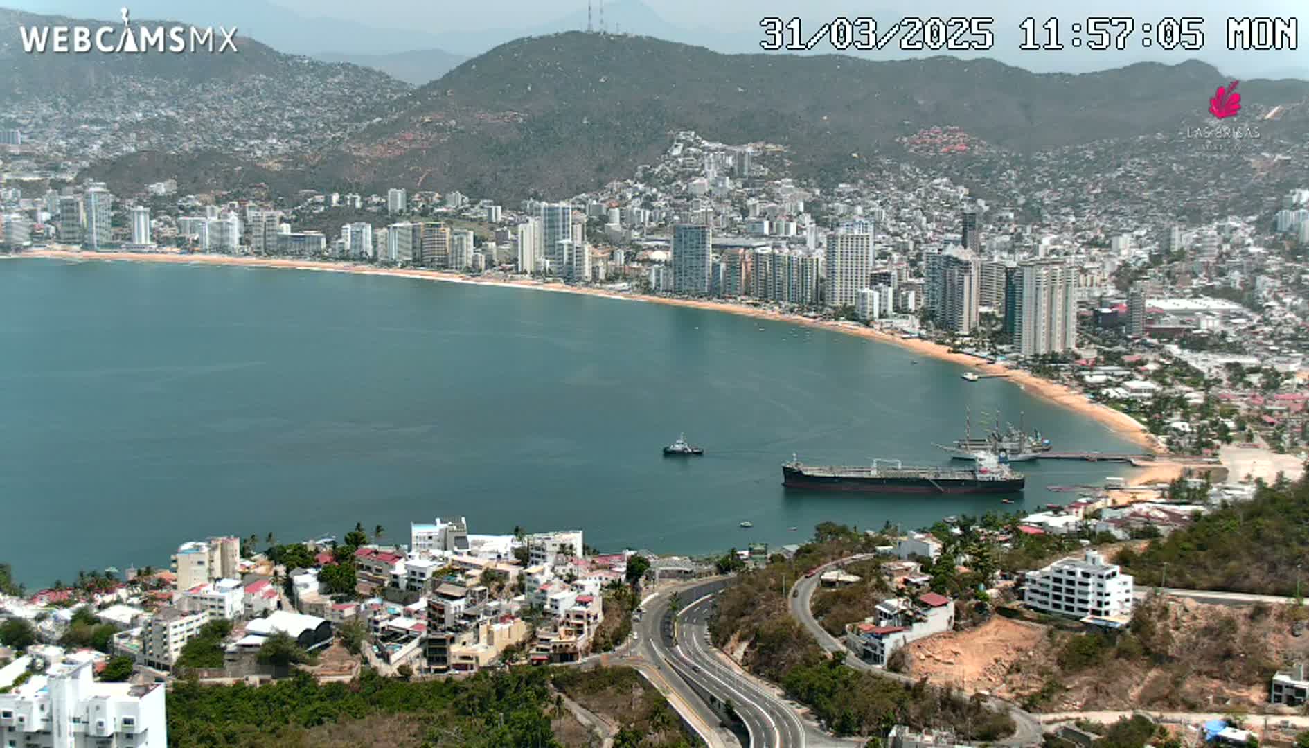 An aerial view shows a coastal city on a partly sunny day, with a large body of calm water curving around a sandy beach lined with high-rise buildings and a large tanker ship in the bay.