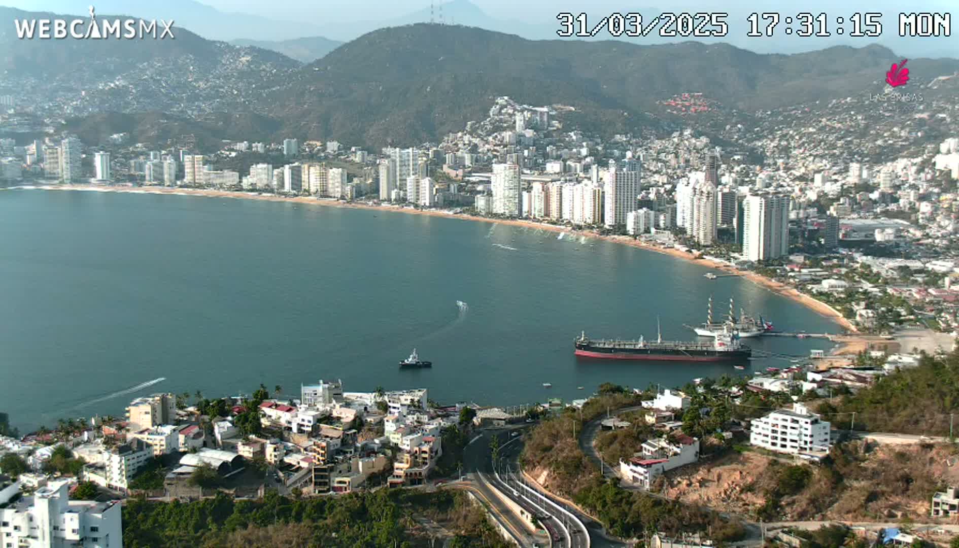An aerial view shows a coastal city with high-rise buildings along a curved bay under a clear sky, with several ships in the harbor.