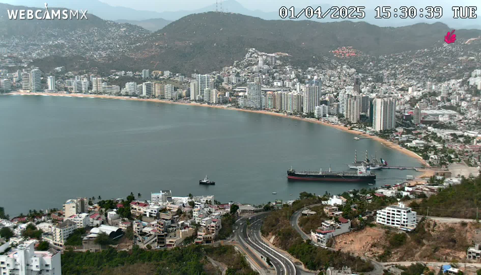 A coastal city with numerous high-rise buildings stretches along a calm bay under a clear sky, with a large tanker and smaller vessel visible in the water.