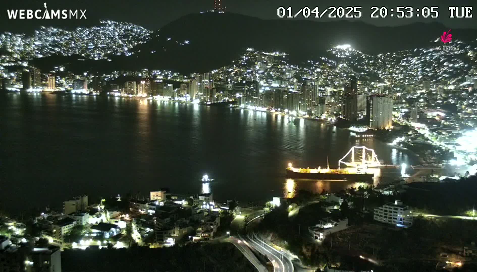 A nighttime aerial view of a city sprawling along a dark bay, with a large ship docked near the shore and the city lights illuminating the hillsides.