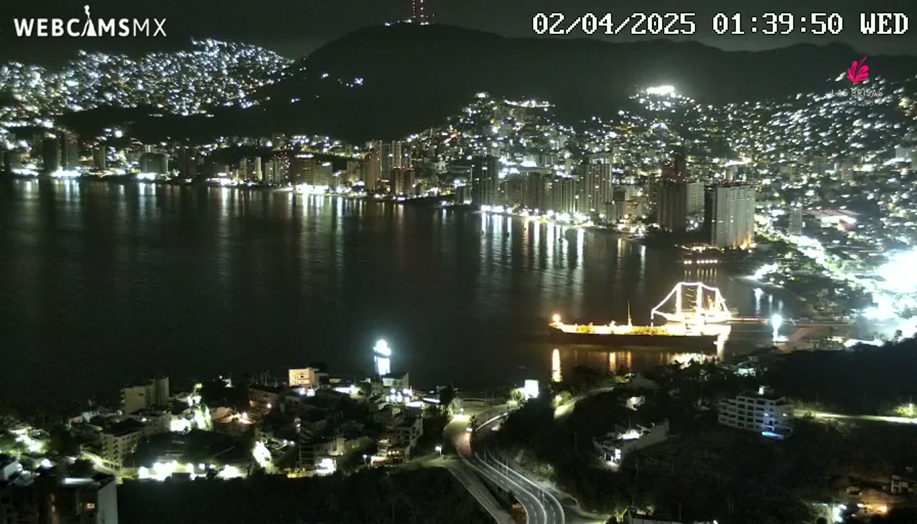 A nighttime view of a city lit up along a calm bay, with a large ship docked near the shore.