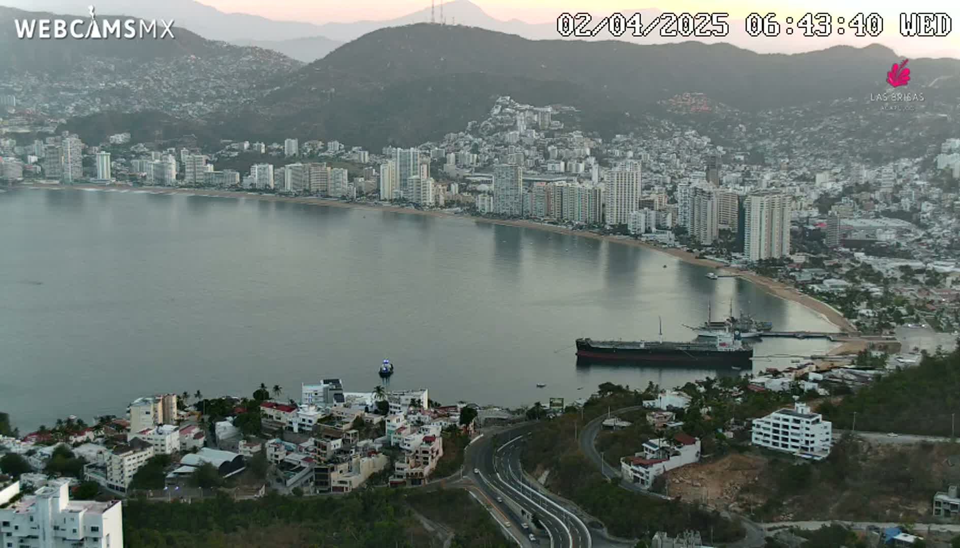 A coastal city with many high-rise buildings sits on a calm bay under a mostly clear sky at dawn, with a large ship docked near the shore and a winding road visible in the foreground.