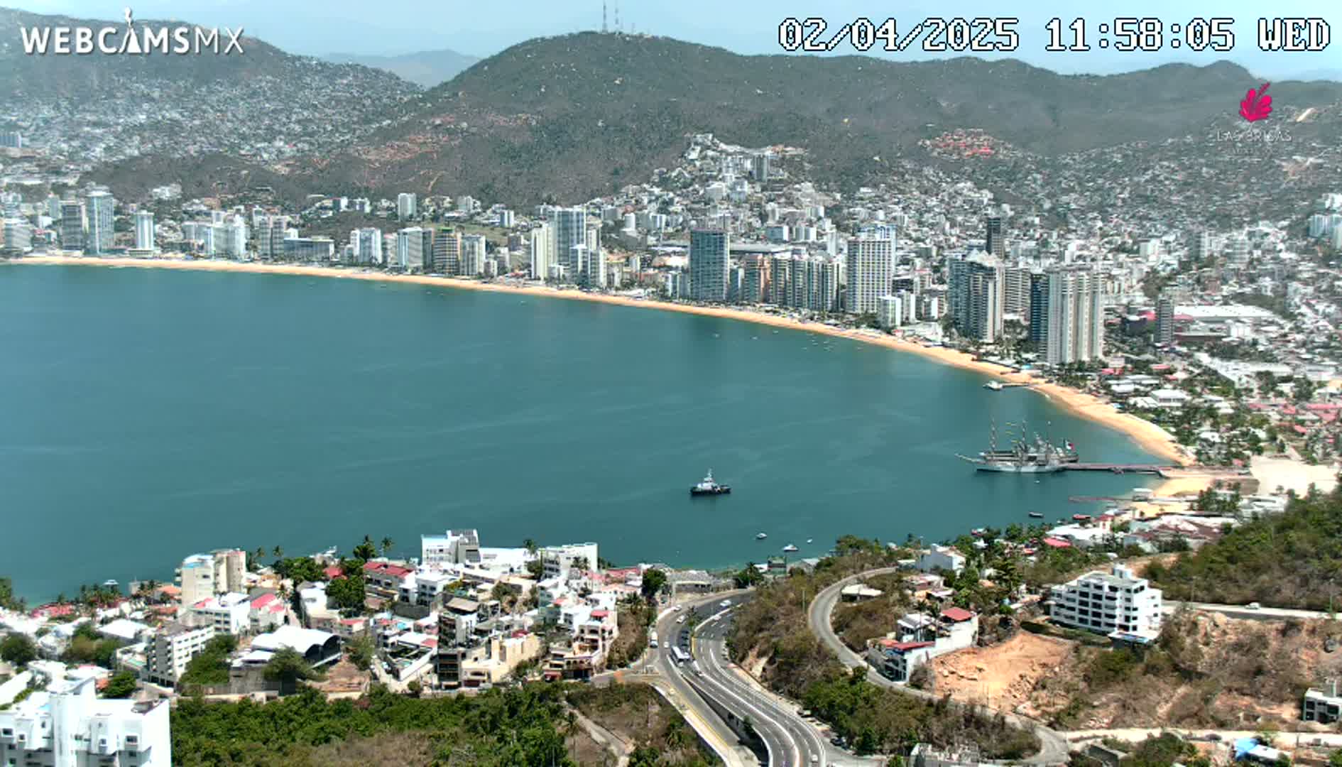 A coastal city with numerous high-rise buildings sprawls along a curved bay under a clear, sunny sky.