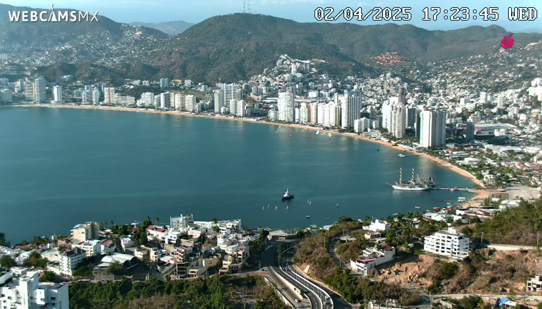 A coastal city with high-rise buildings and a sandy beach curves around a calm bay under a clear sky.
