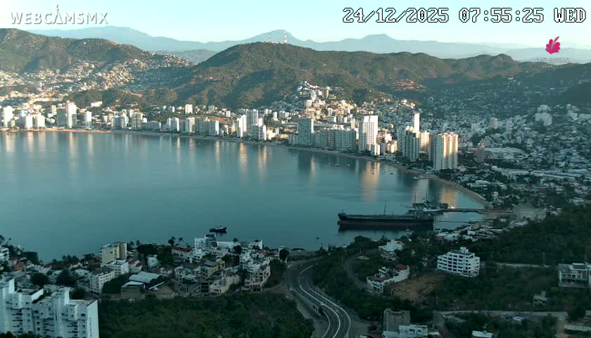 A wide panoramic view of a vibrant city bay at night features countless lights from buildings and residences reflecting on the calm water, with a large cargo ship and a smaller boat visible, all under a clear sky.