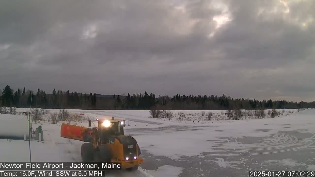 A yellow snowplow clears a snow-covered, partially frozen airfield under an overcast sky.