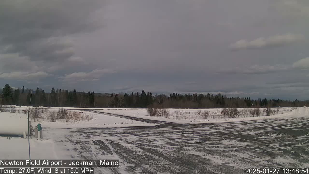 A snow-covered airfield with a line of evergreen trees in the background under a mostly cloudy sky.