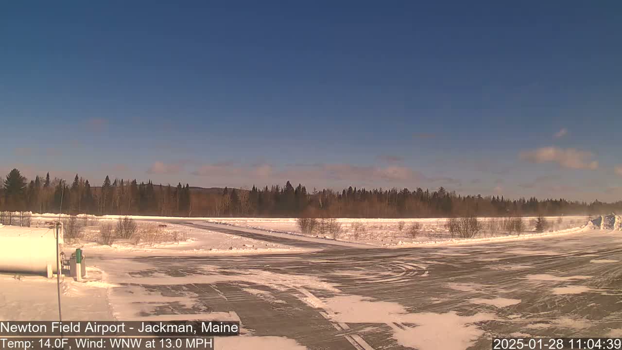 A snow-covered airport tarmac extends to a line of trees under a clear blue sky.