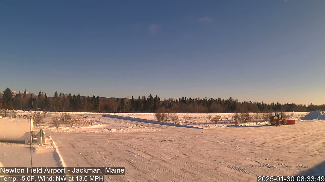 A snow-covered airfield with a snowplow, under a clear blue sky, and a background of snow-covered evergreen trees.