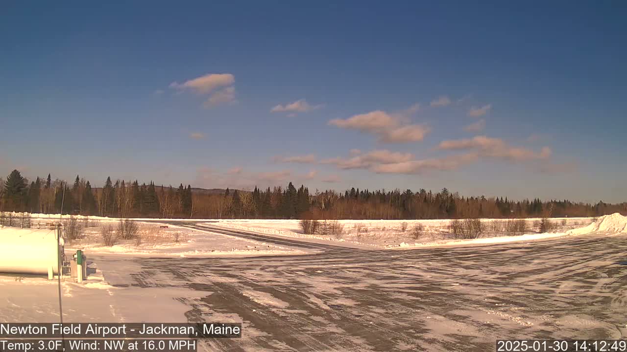 A snow-covered airfield is visible under a mostly clear, bright blue sky with a few scattered clouds, and a line of trees borders the airfield in the background.