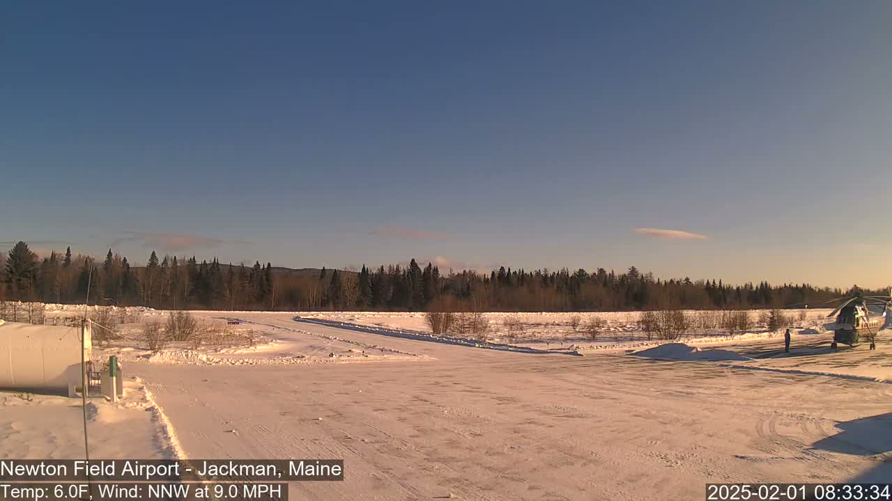 A snow-covered airfield with a helicopter and a line of trees in the background under a clear, sunny sky.