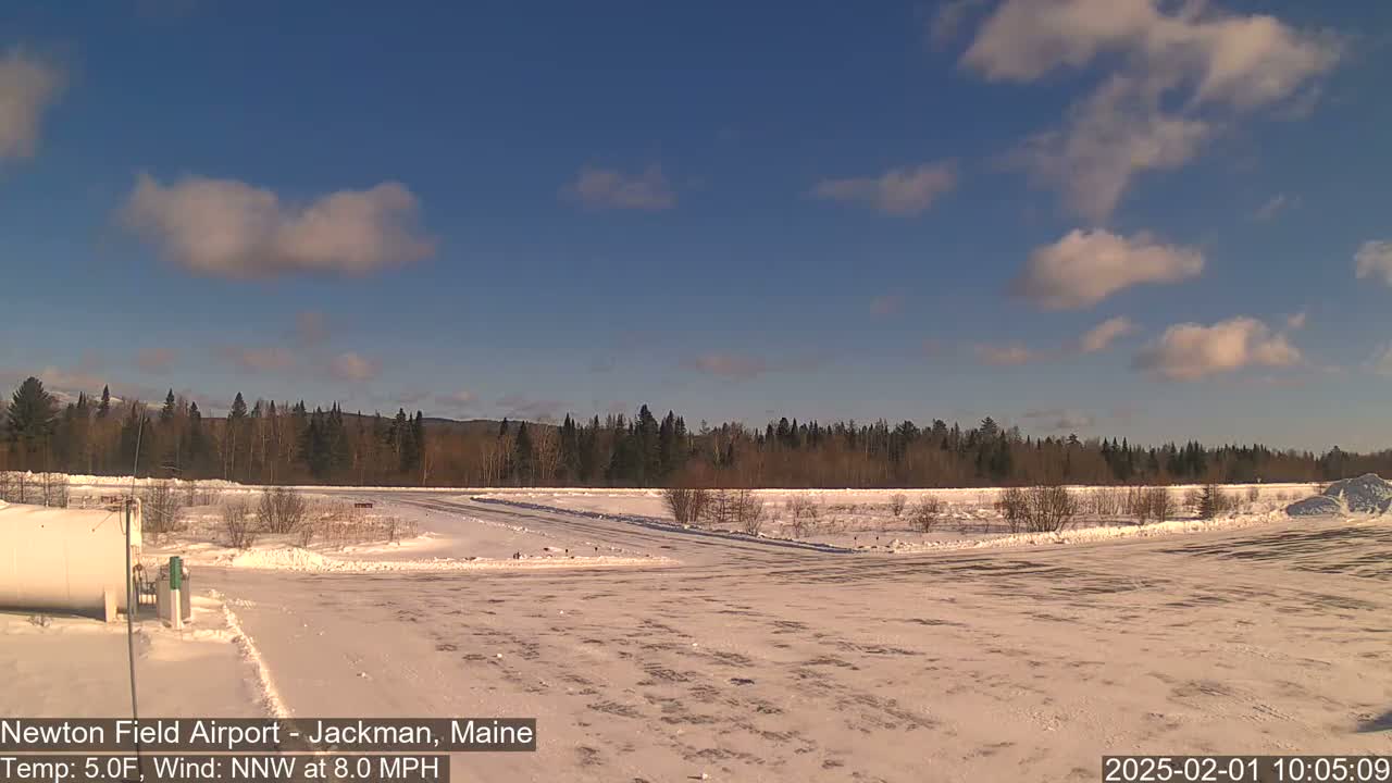 A snow-covered airfield is visible, with a line of trees in the background under a mostly clear, bright blue sky with a few scattered clouds.