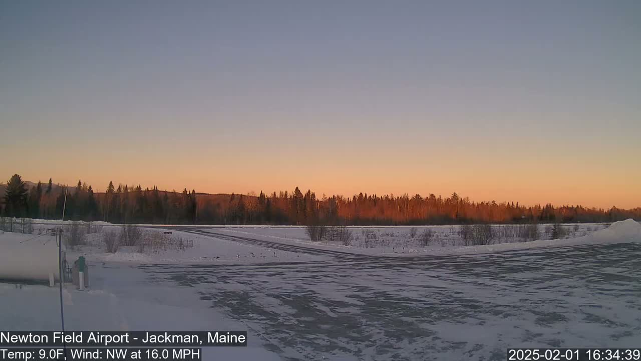 A snow-covered airfield at sunset, with a line of dark trees in the background under a pale orange and blue sky, and a northwest wind blowing at 16 mph.