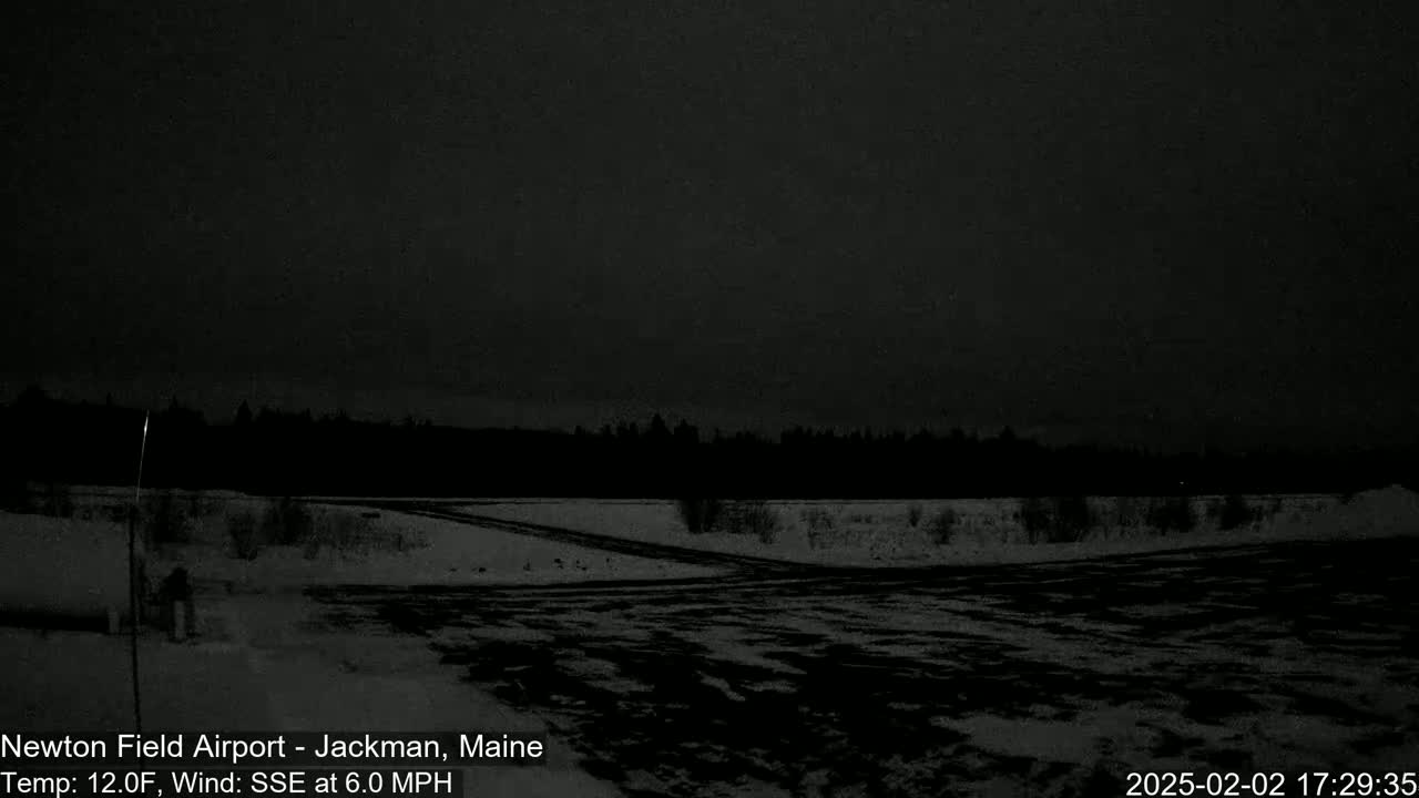 A dark, snowy, outdoor scene at night shows a mostly empty snow-covered parking lot and road, with a dark treeline in the background under a dark sky.