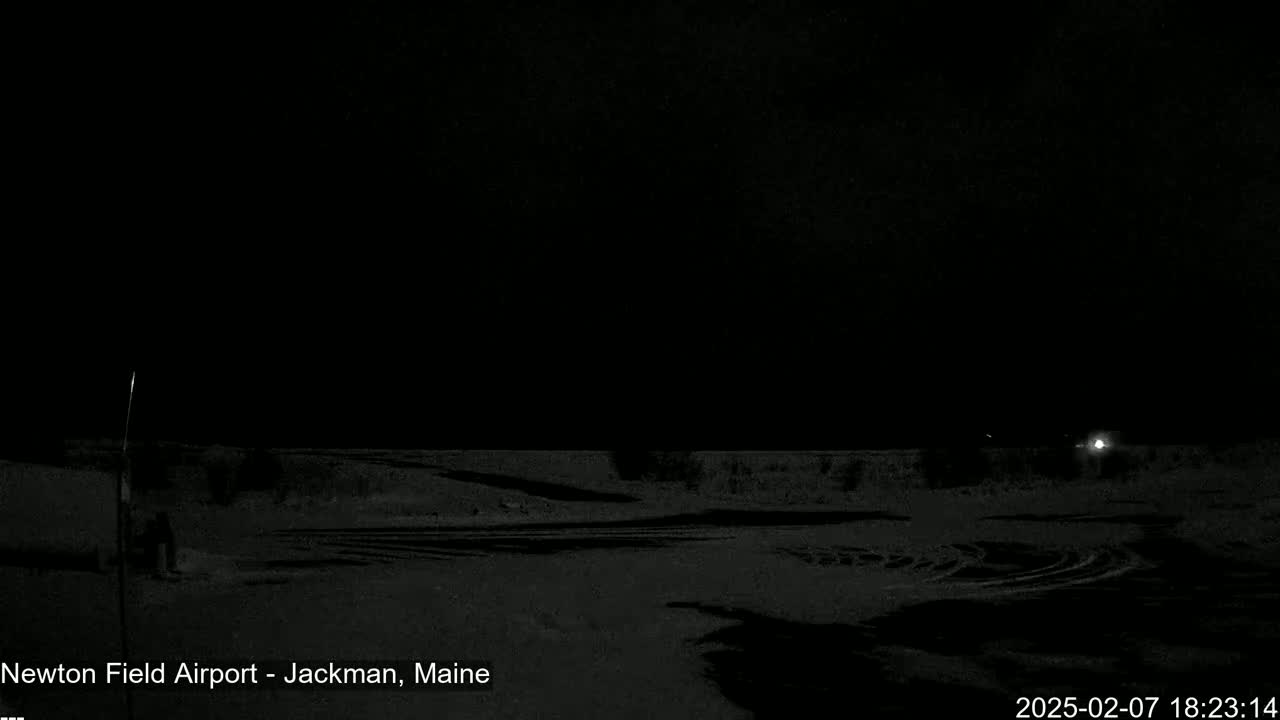 A dark, snowy, nighttime scene shows a flat expanse of land with tire tracks under a black sky.