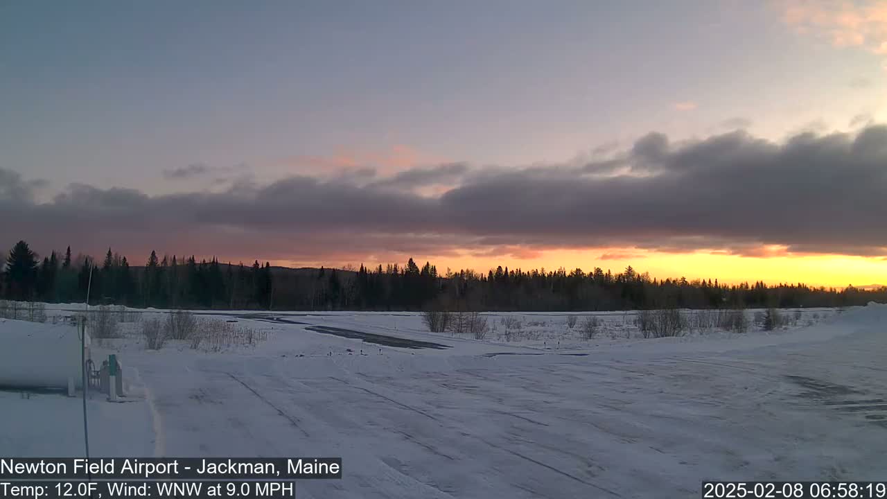 A snow-covered airfield at sunset, under a partly cloudy sky, with a line of dark trees in the background.