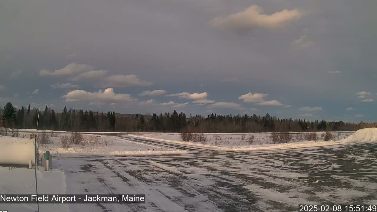 A snow-covered airport runway stretches toward a line of trees under a partly cloudy sky.