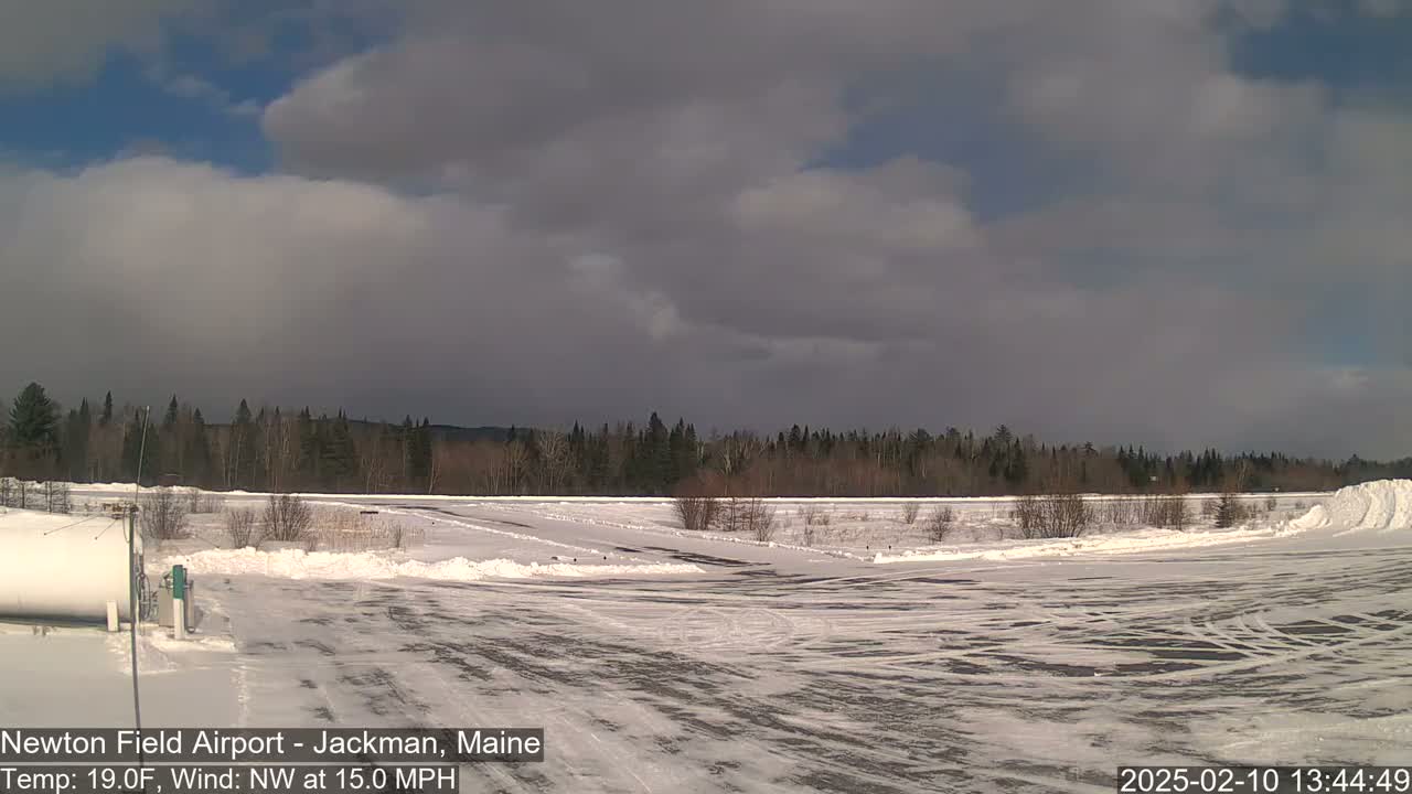 A snow-covered airfield with a line of trees in the background under a partly cloudy sky with a northwest wind at 15 mph.