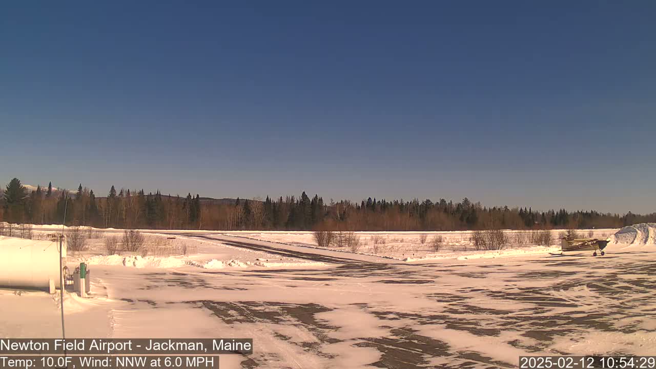 A snow-covered airfield with a small airplane parked near a snowbank, under a clear blue sky.