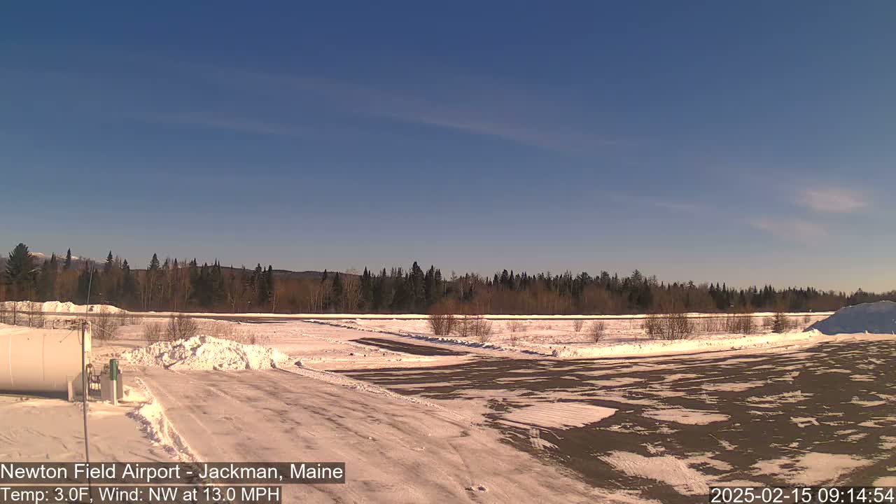 A snow-covered airfield with a dark-colored paved area, bordered by a line of trees under a clear blue sky.
