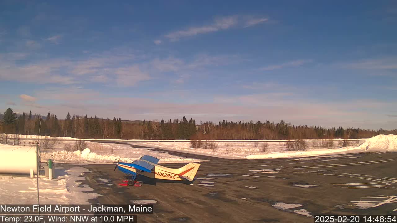 A small airplane sits on a snow-covered airfield under a mostly clear, bright blue sky.