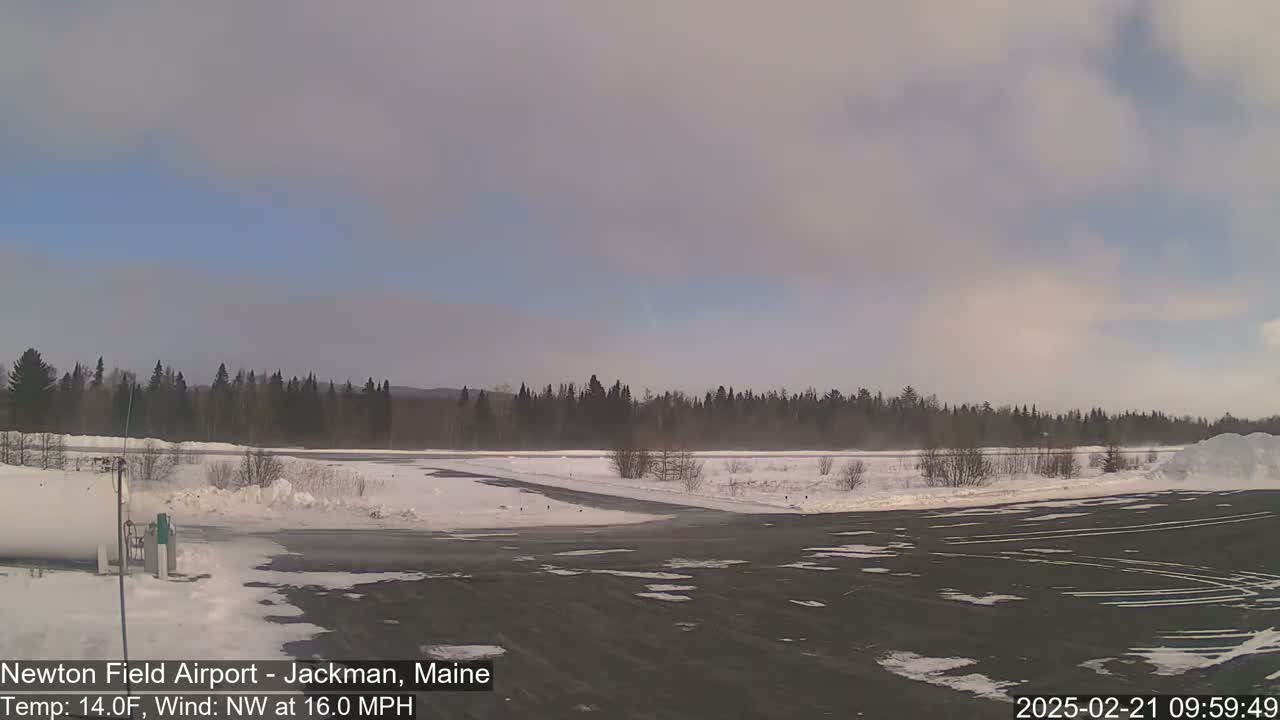 A snow-covered airfield with a dark asphalt runway and a line of evergreen trees in the background under a partly cloudy sky.