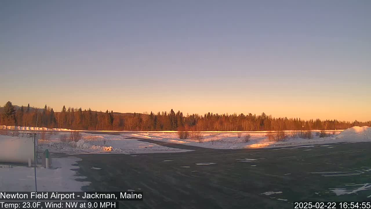 A snow-covered airfield at sunset, with a line of trees in the background under a clear, pale orange sky and a northwest wind.