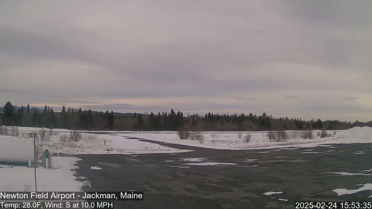 A snow-covered airfield with a dark asphalt runway is bordered by a line of evergreens under an overcast sky.