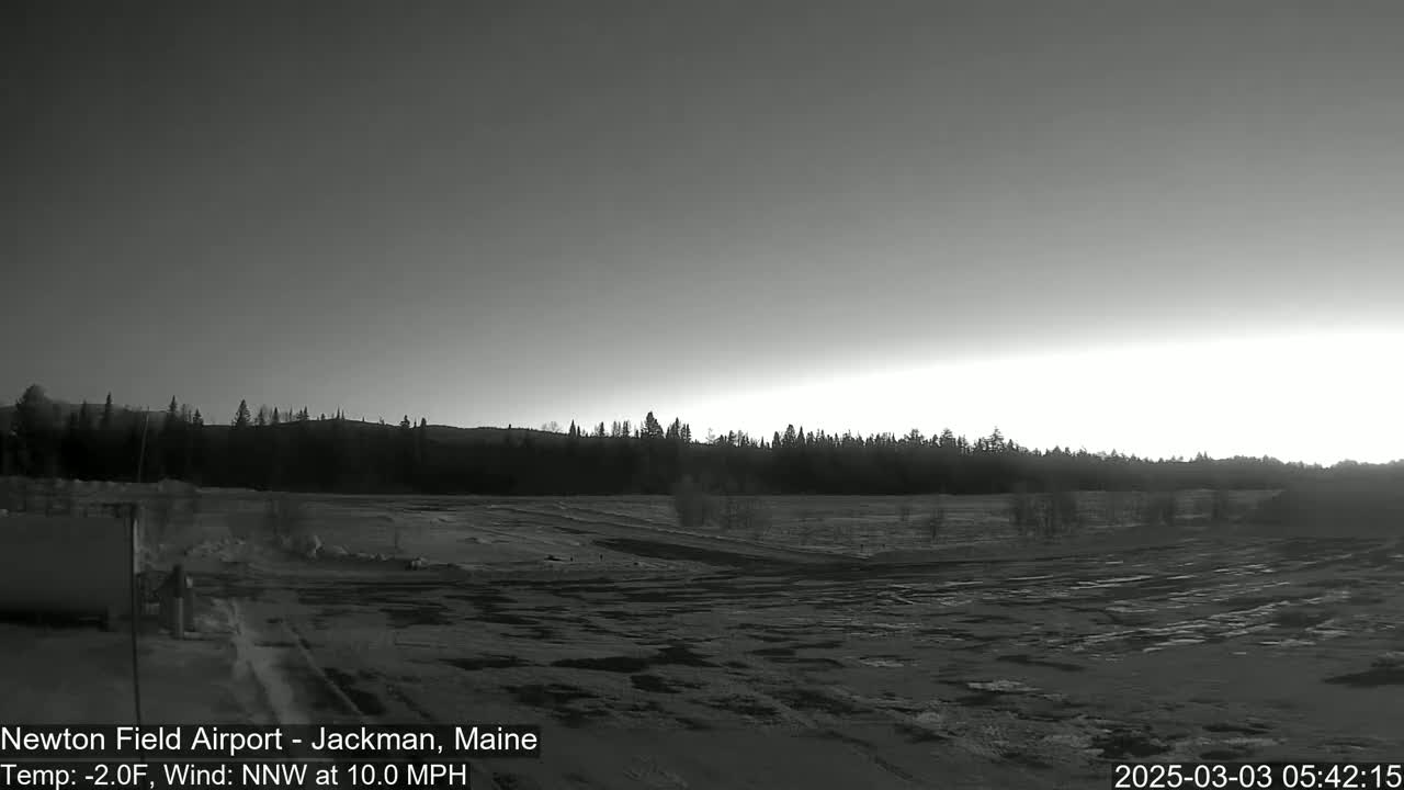 A snow-covered airfield is visible at dawn under a mostly clear sky, with a line of trees in the background and a light wind.