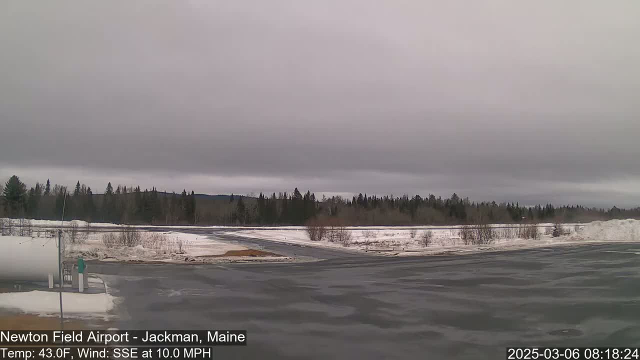 A mostly overcast sky hangs over a snow-covered airfield with a line of dark evergreen trees in the background.