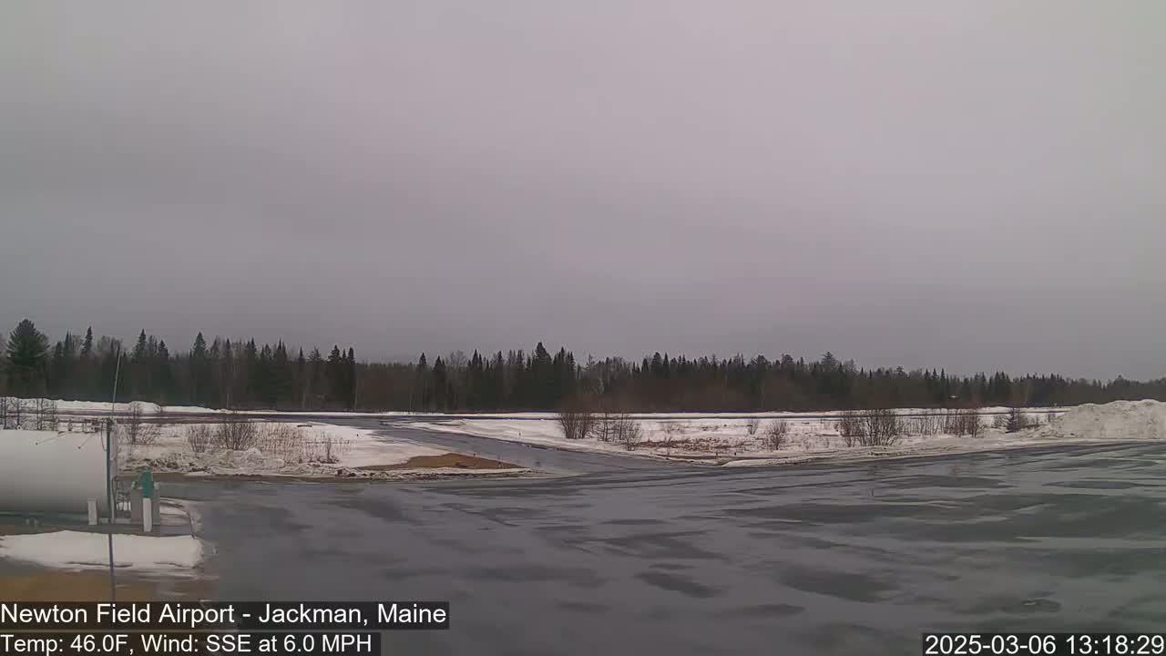 A mostly overcast day shows a snow-covered airfield with a line of dark evergreens in the background.