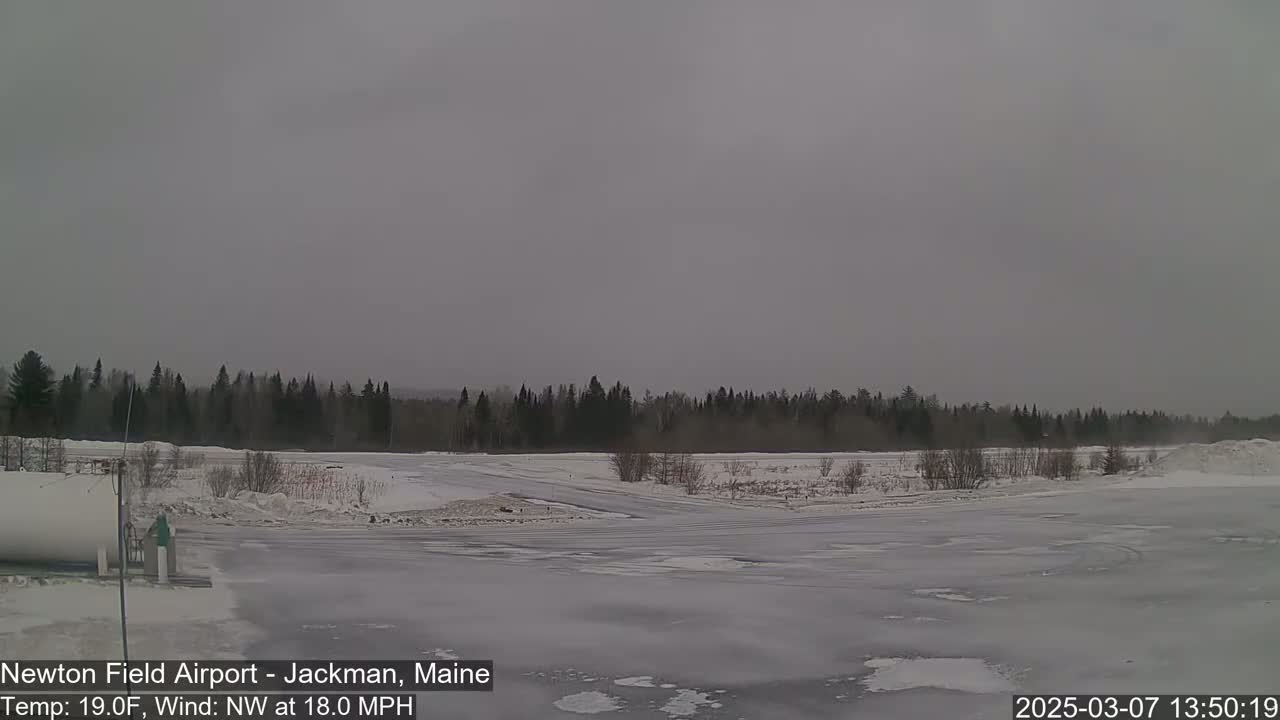 A snow-covered, icy expanse of land stretches before a line of dark evergreens under an overcast sky, with an 18 mph northwest wind.