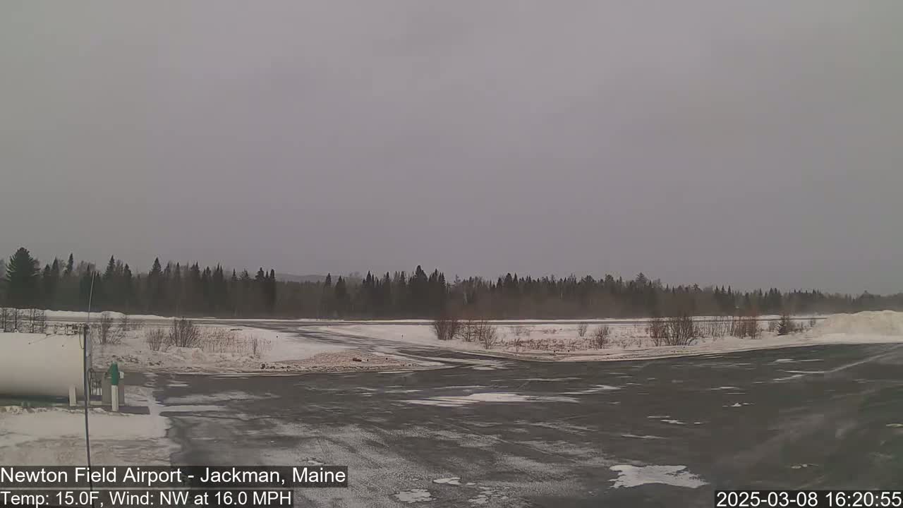 A snow-covered airfield with a dark asphalt runway is visible, under an overcast sky with a northwest wind.