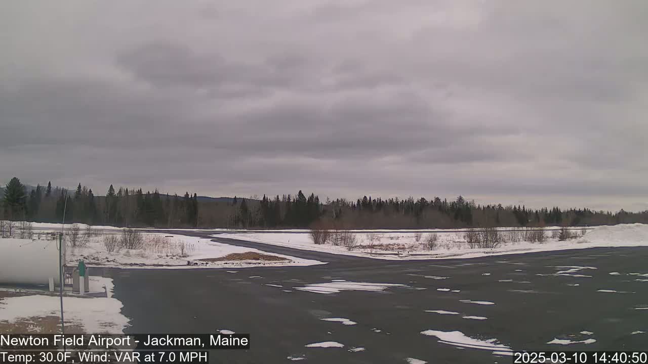 A mostly overcast sky hangs over a snow-covered airport tarmac and surrounding forest.