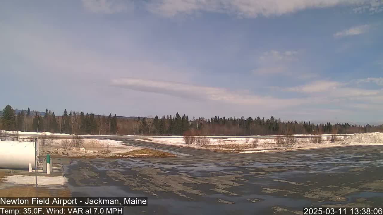 A mostly sunny day shows a snow-dusted airport tarmac and runway bordered by a line of dark evergreen trees under a partly cloudy sky.