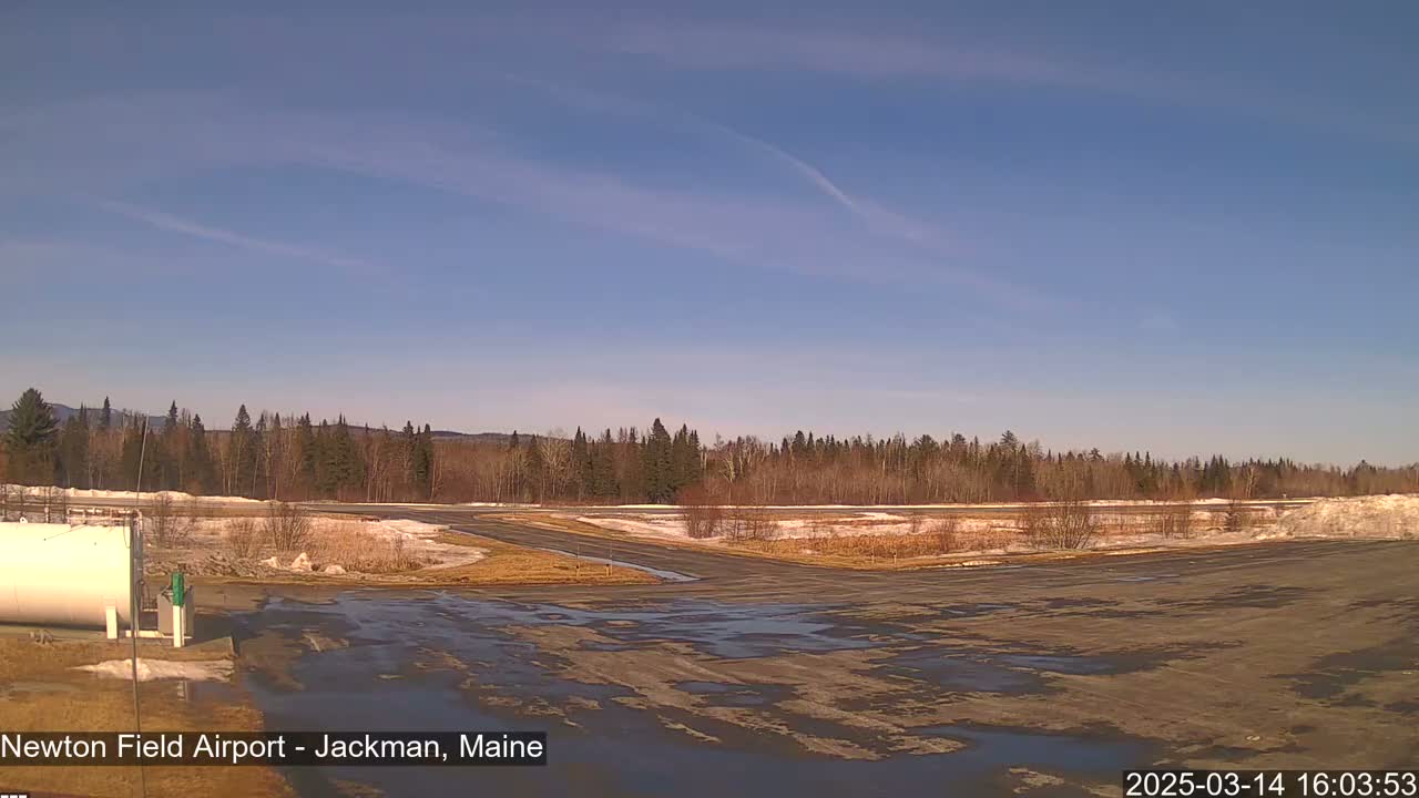 A mostly clear, sunny day reveals a snow-dusted airfield bordered by a line of bare deciduous trees and evergreen conifers, with patches of melted snow and ice on the ground.