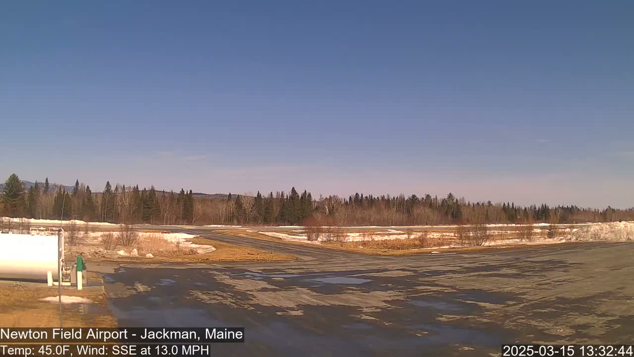 A mostly clear, sunny day at an airfield shows a tarmac with patches of melting snow and ice, bordered by trees and distant mountains.