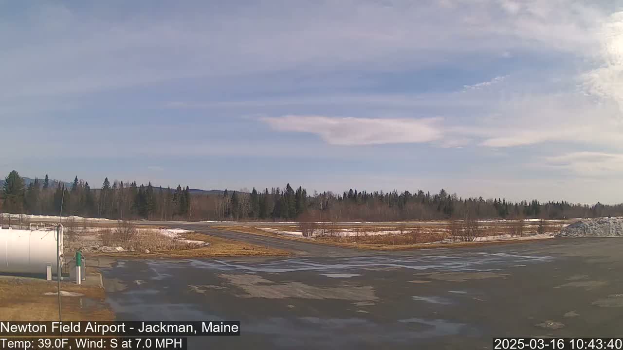 A mostly sunny airfield with a few clouds, a smattering of snow on the ground, and trees in the background.