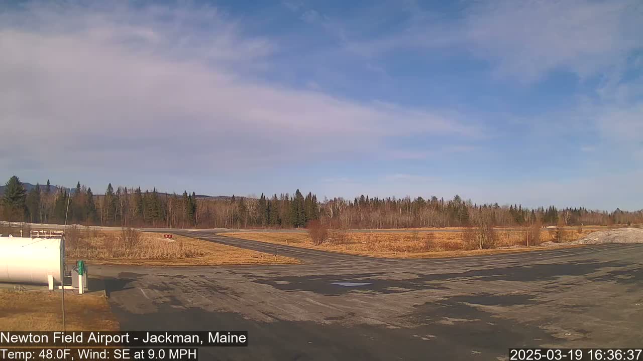 A small airport runway with a few buildings and a large expanse of trees and brown fields under a mostly clear, blue sky.