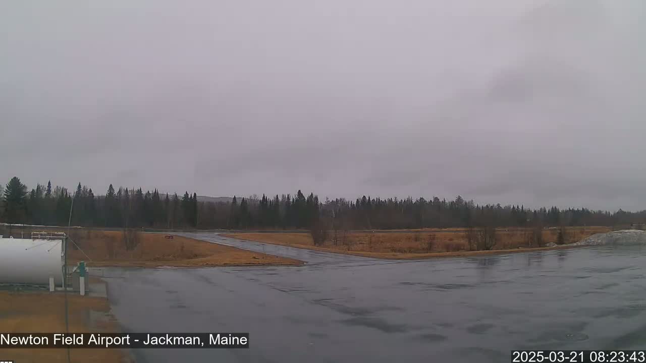 A wet, overcast day at an airport with a large fuel tank and a runway leading to a line of trees.