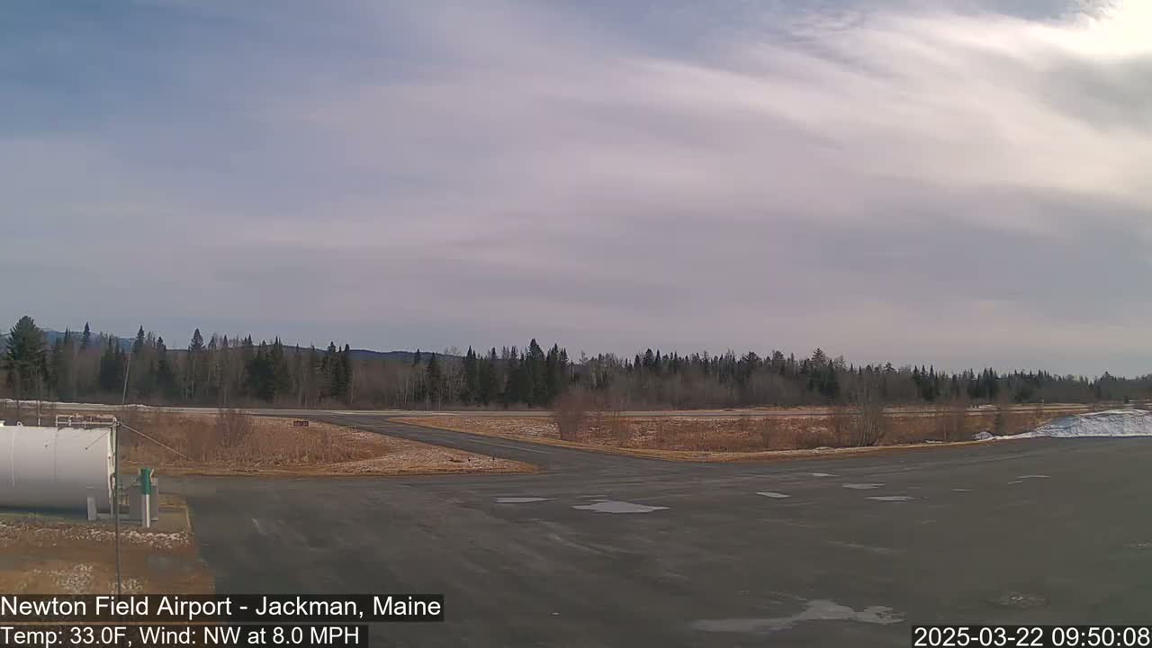 A mostly cloudy day at an airfield shows a paved area with a fuel tank, leading to a grassy field bordered by a line of trees under a pale sky.