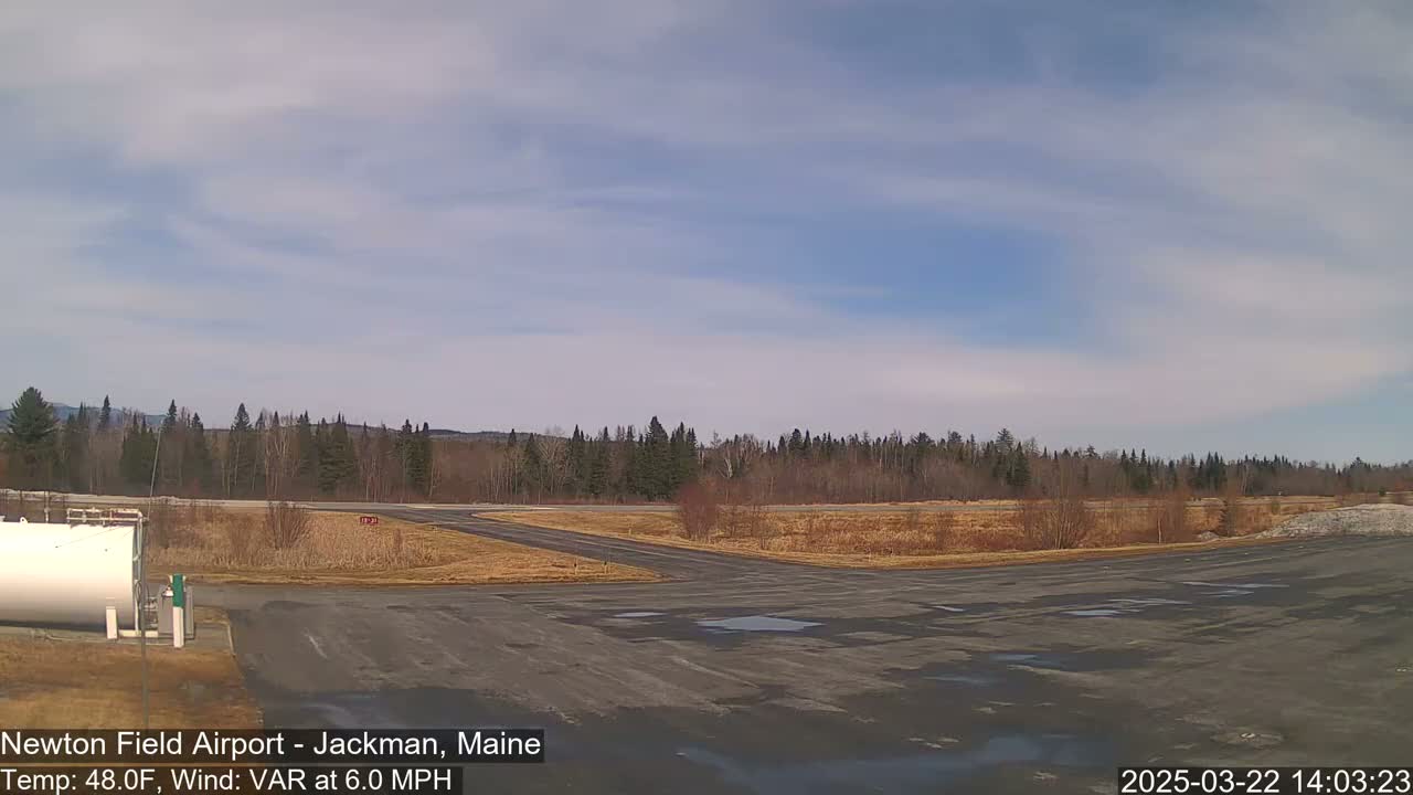 A mostly sunny day shows a small airport tarmac with a line of trees in the background and a few puddles on the ground.