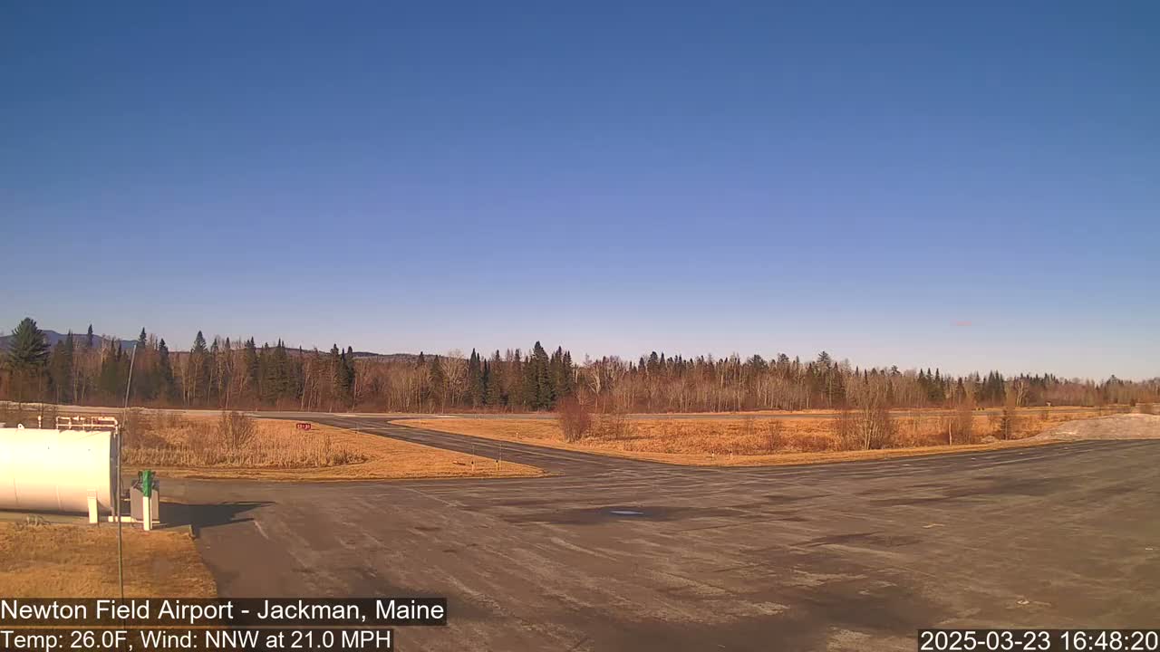 A small airport tarmac with a line of trees in the background under a clear blue sky and a 26°F temperature with a 21 mph north-northwest wind.