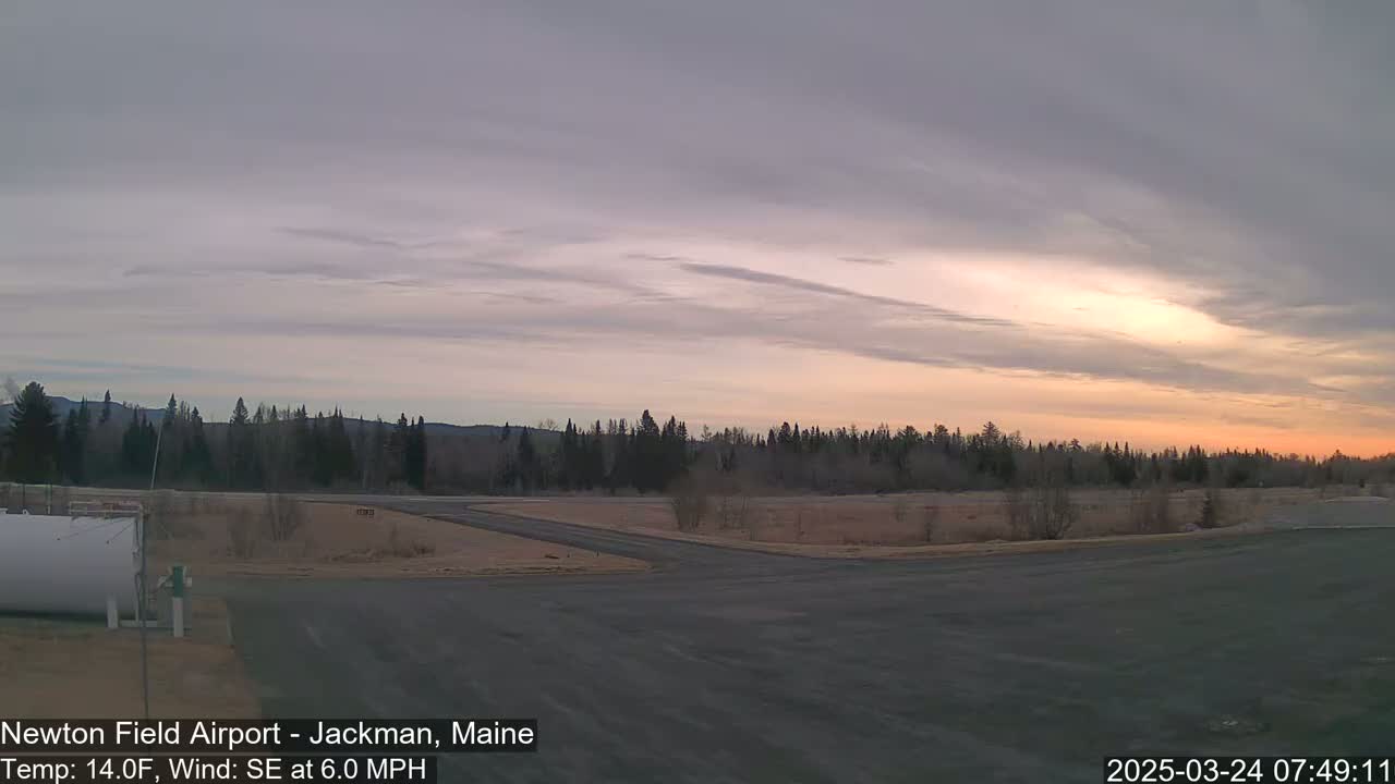 A mostly cloudy sunrise illuminates a flat, sparsely vegetated airfield with a line of trees in the background.