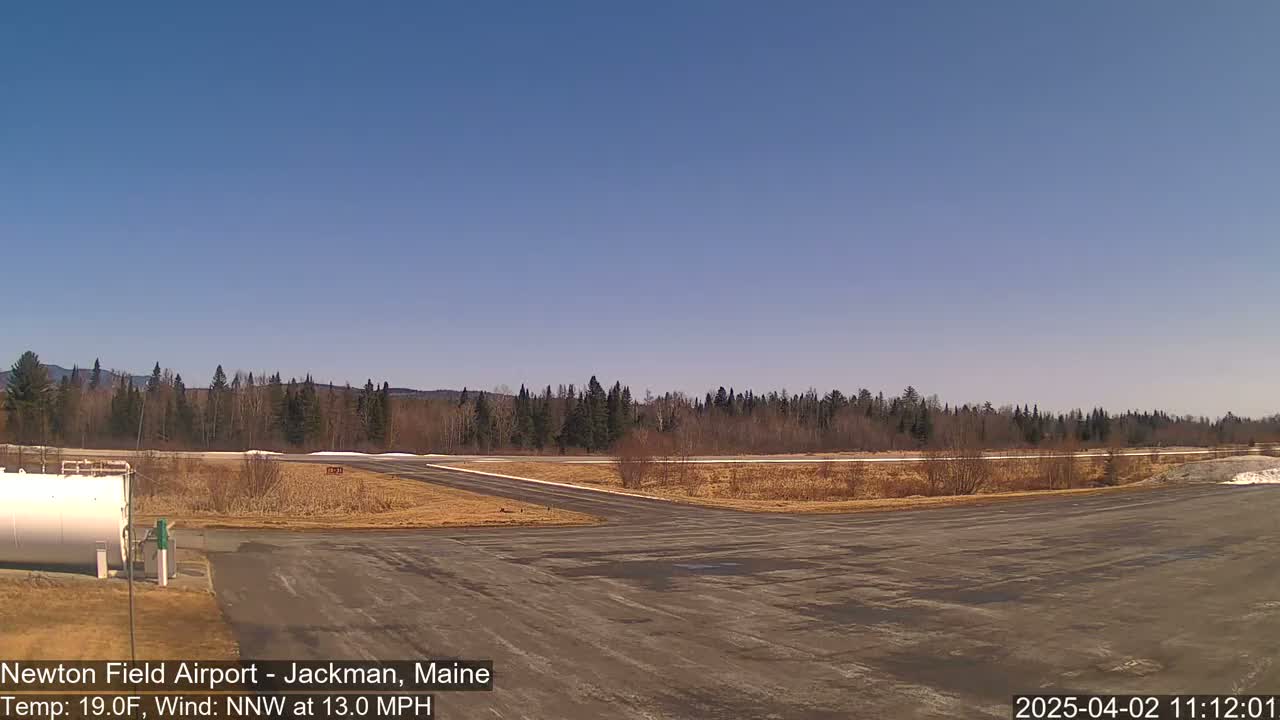 A mostly clear, sunny day with a north-northwest wind at 13 mph shows an airport tarmac with a line of trees and distant mountains in the background.