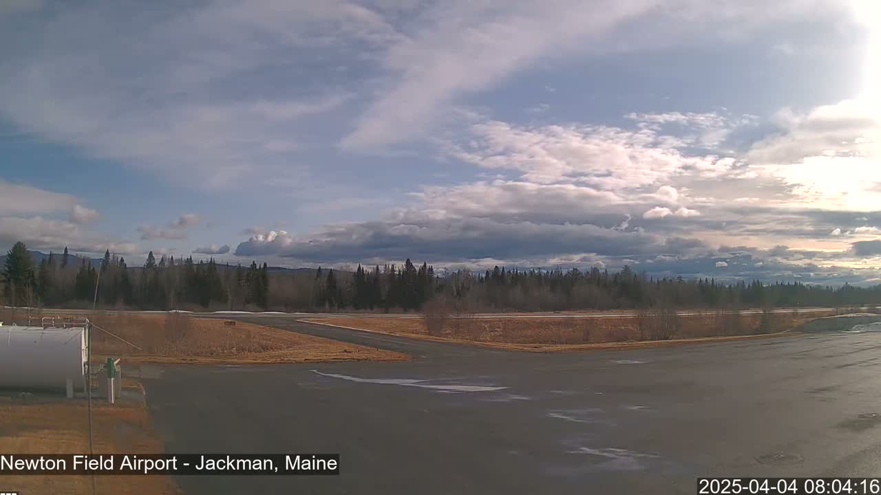 A mostly cloudy sky over a flat, mostly barren airfield with a line of evergreens in the background.