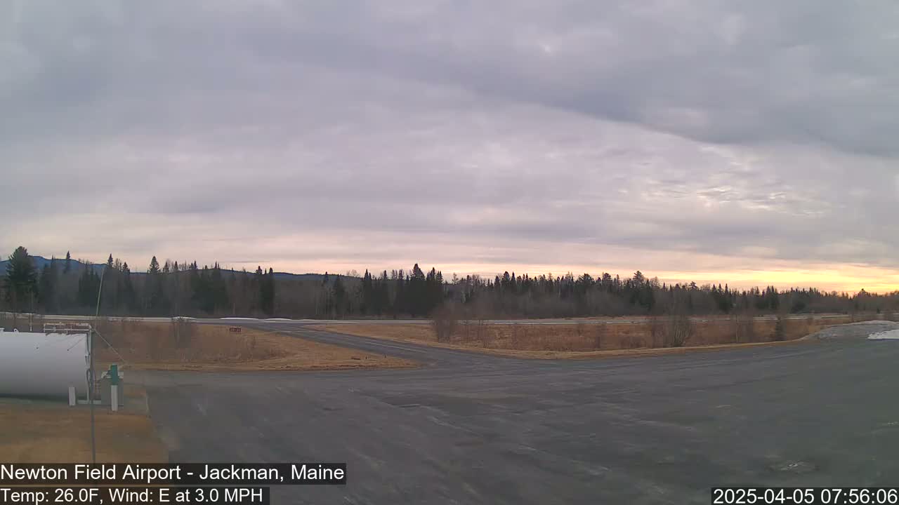 A mostly cloudy sunrise illuminates an airfield with a large, empty tarmac, a white fuel tank, and a line of trees in the background.